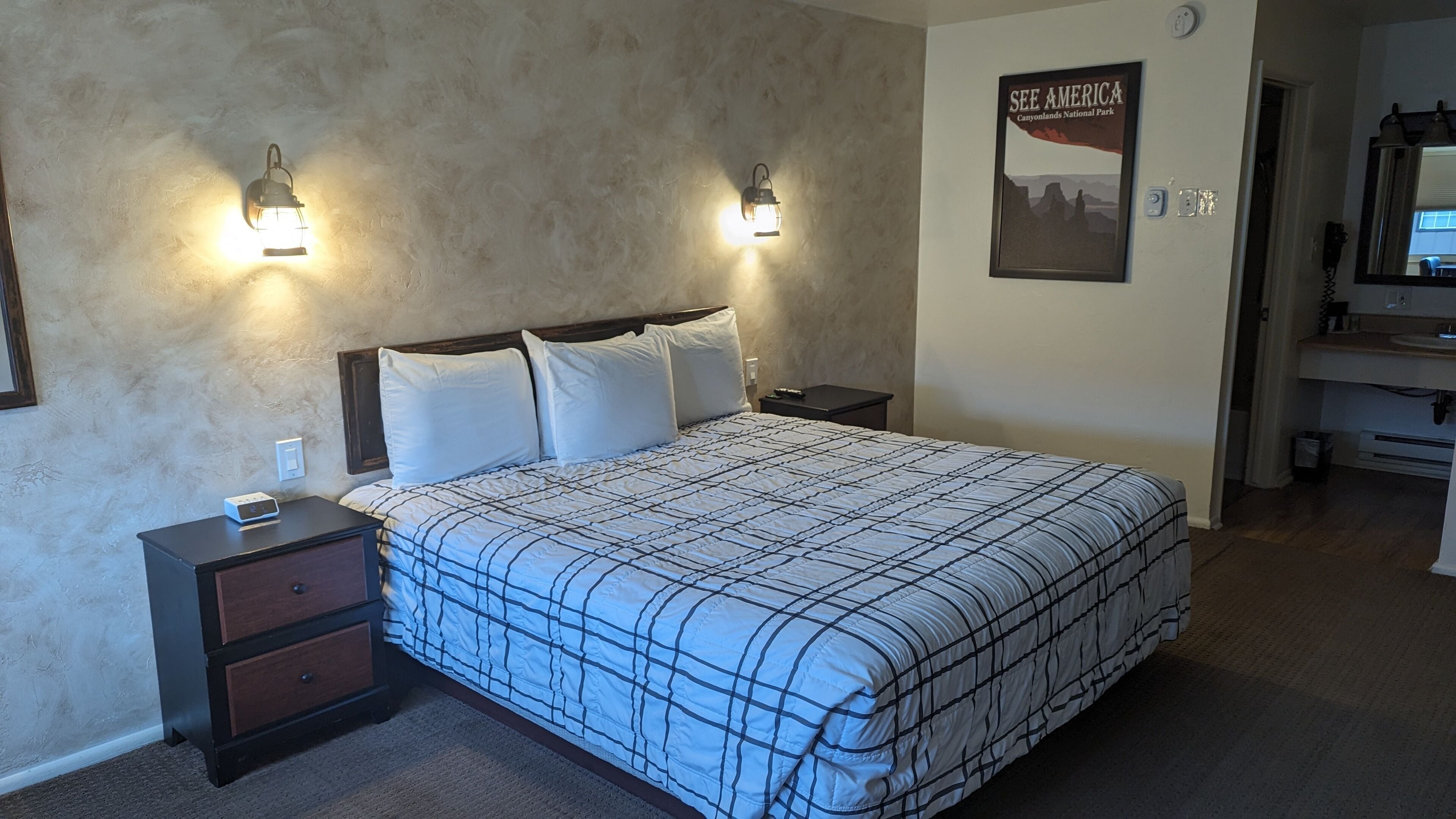 A bedroom with a large bed, two bedside tables, a nightstand, a poster on the wall, and a bathroom visible through an open door.
