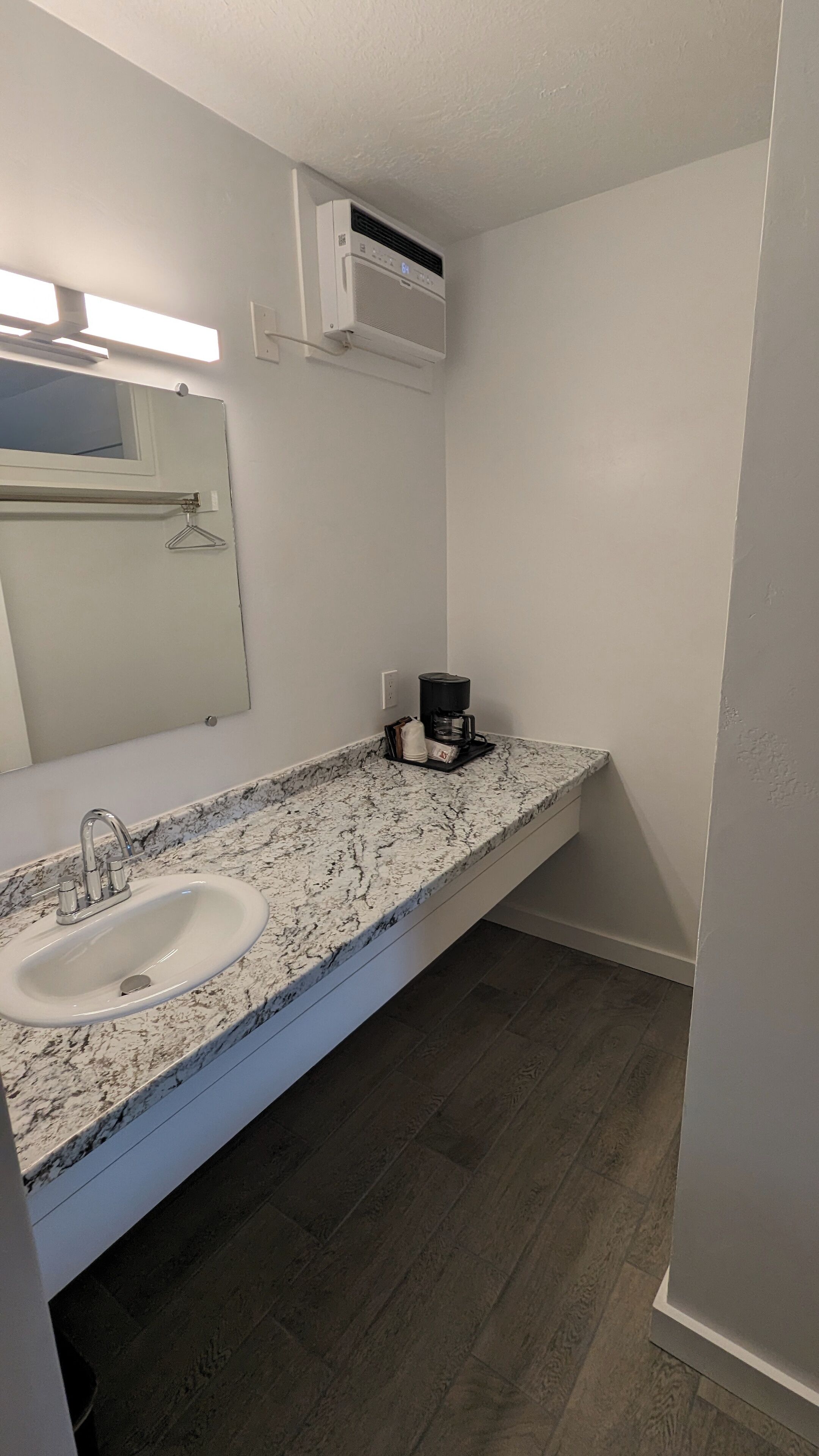 Un baño con lavamanos blanco, un espejo, toallero, cafetera y unidad de aire acondicionado.