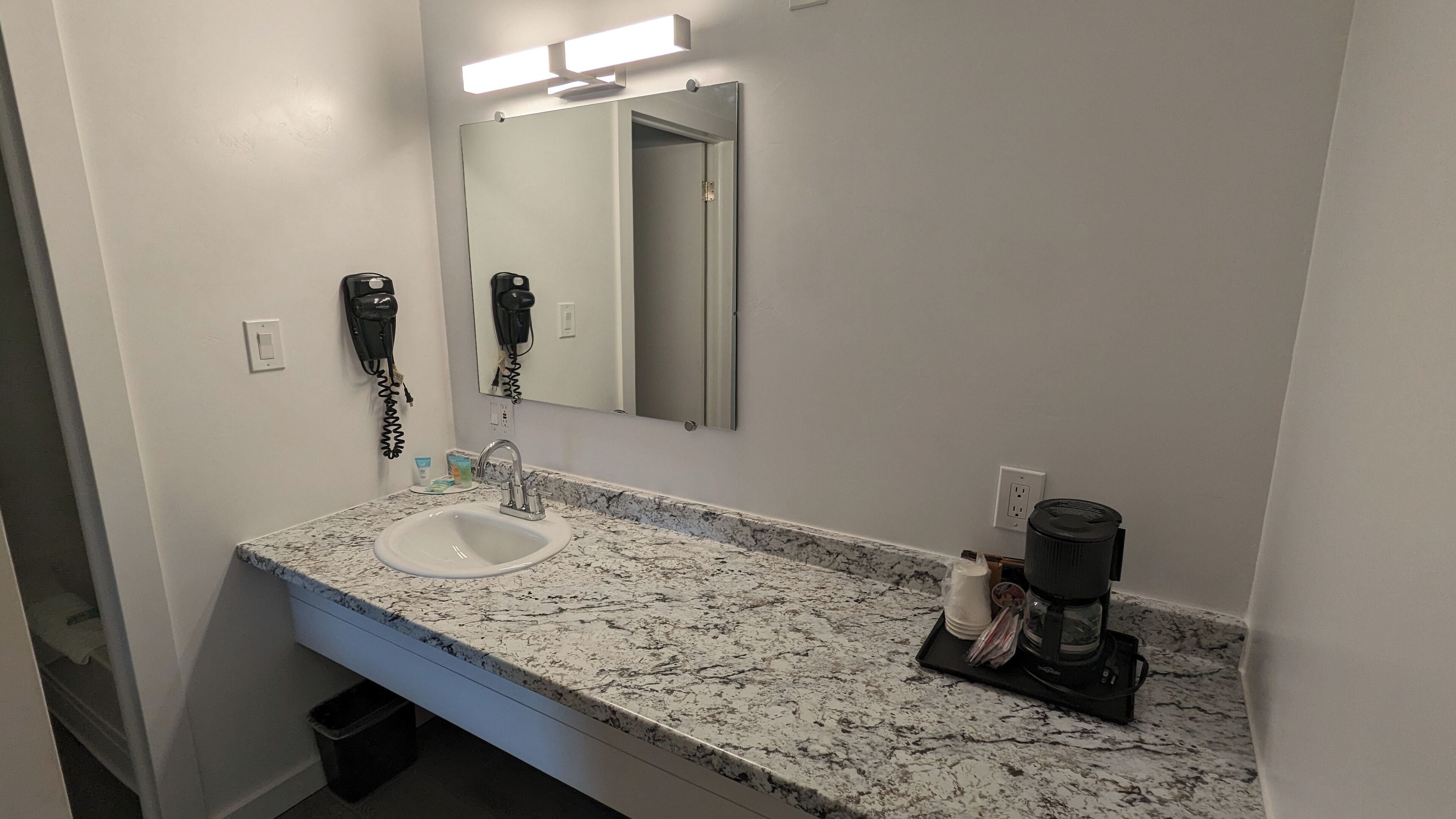 Un baño con encimera de granito, lavamanos, espejo, secador de pelo y cafetera.