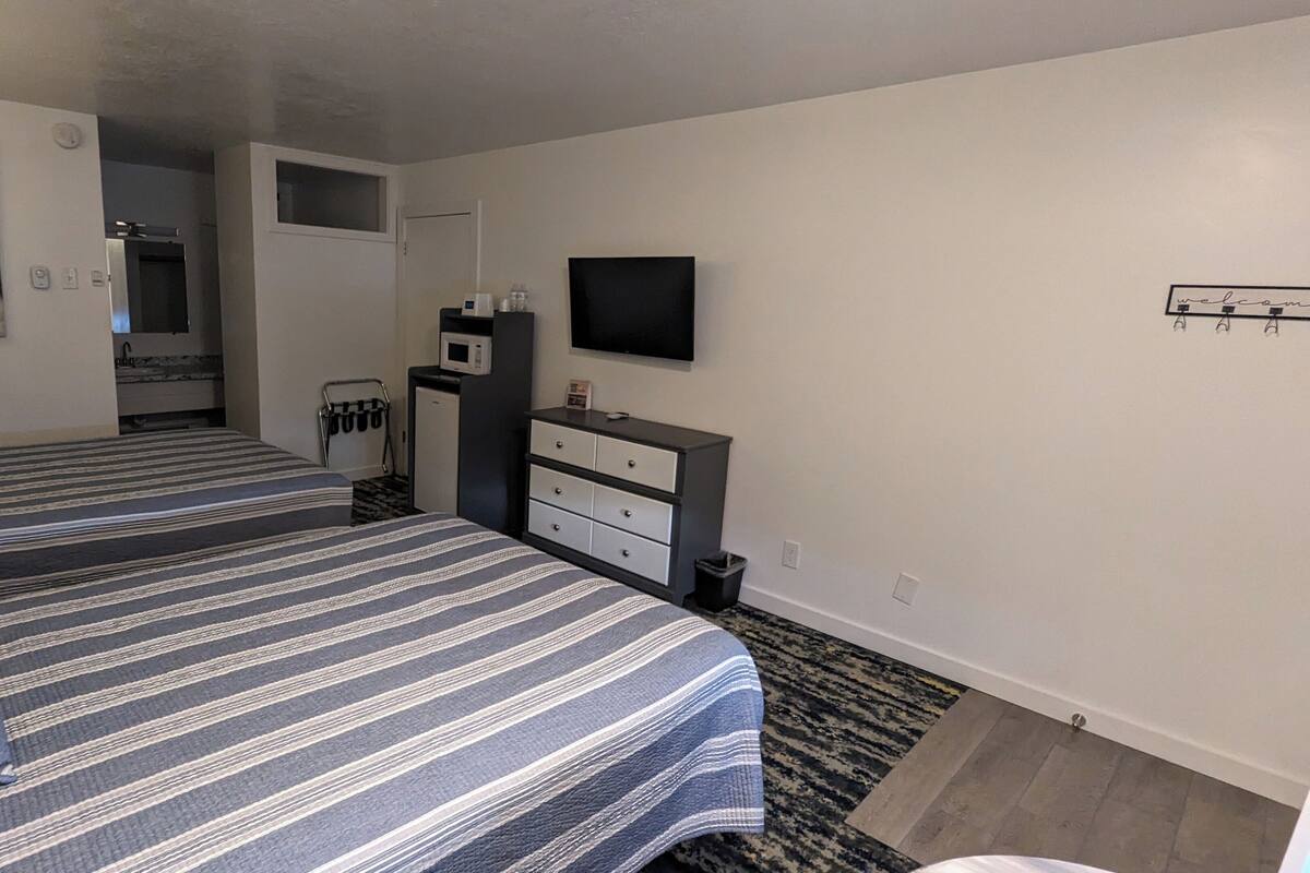 A hotel room with two beds, a TV, a microwave, a small kitchenette, and a painting on the wall.