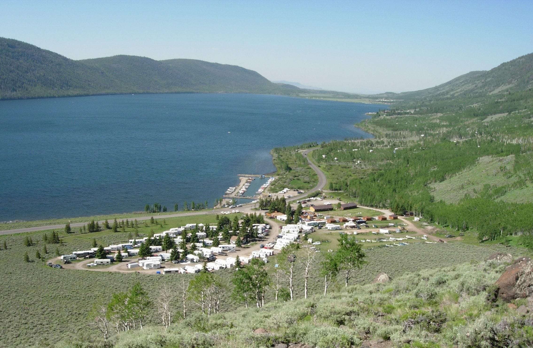 Un camping con vehículos recreativos y tiendas de campaña cerca de un lago, rodeado de colinas.