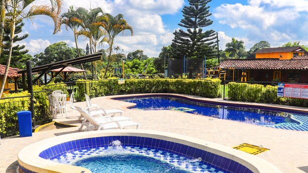 Piscina com espreguiçadeiras e hidromassagem, cercada por vegetação e um edifício com telhado telhado.