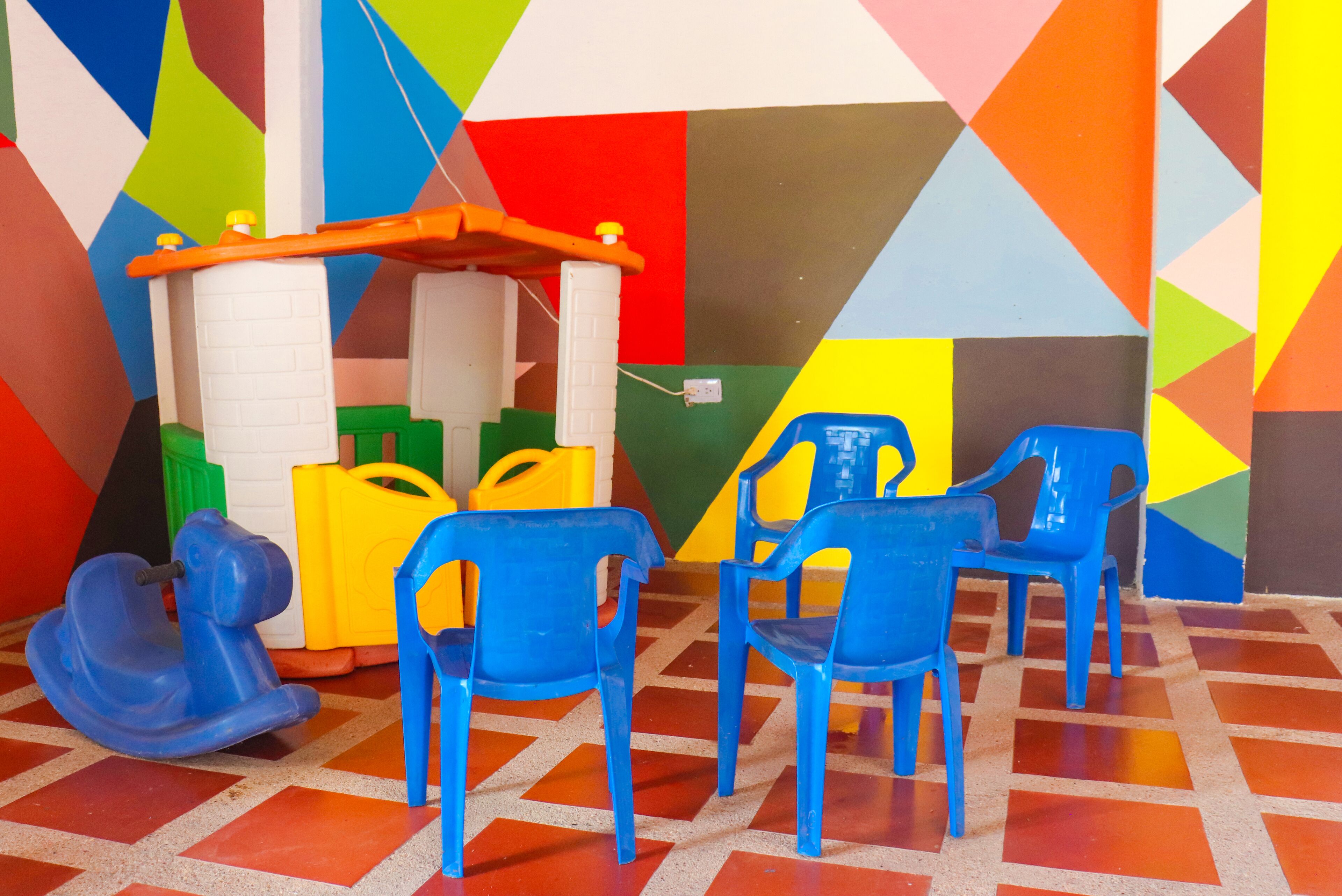 Un espacio de juego colorido con diseños geométricos en la pared, un caballo balanc ín y sillas de plástico azules.