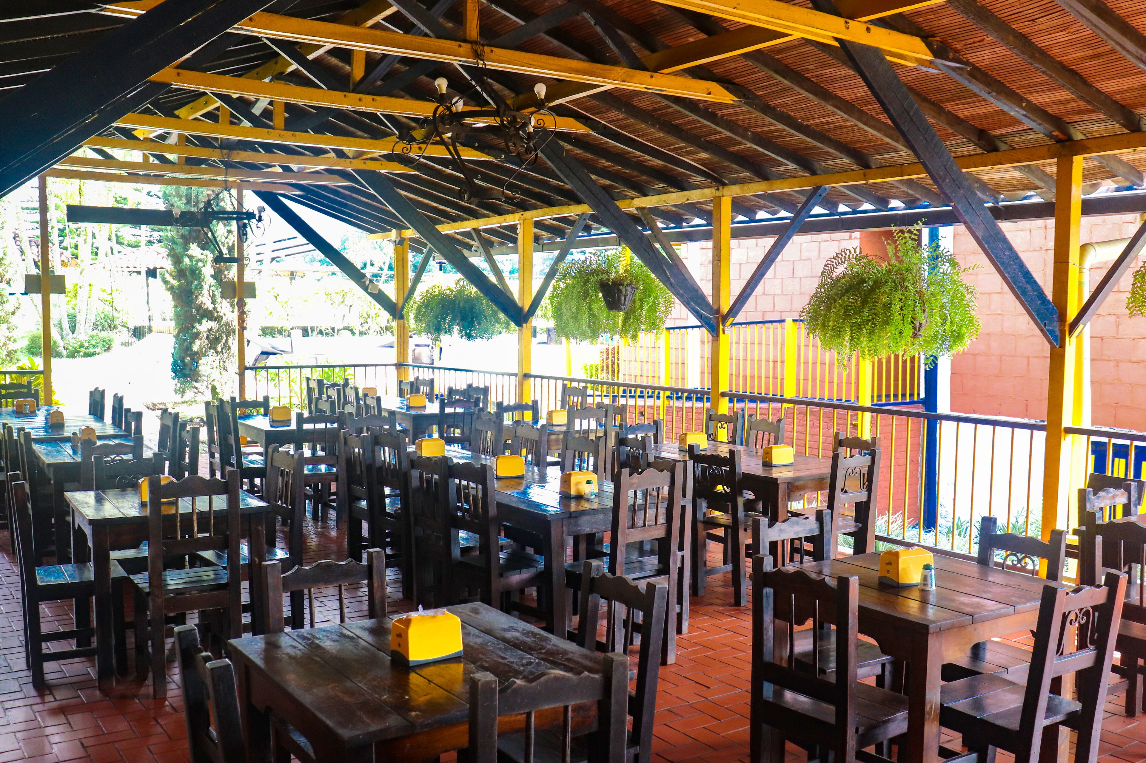 A covered outdoor dining area with wooden tables and chairs, a tiled floor, and hanging plants.