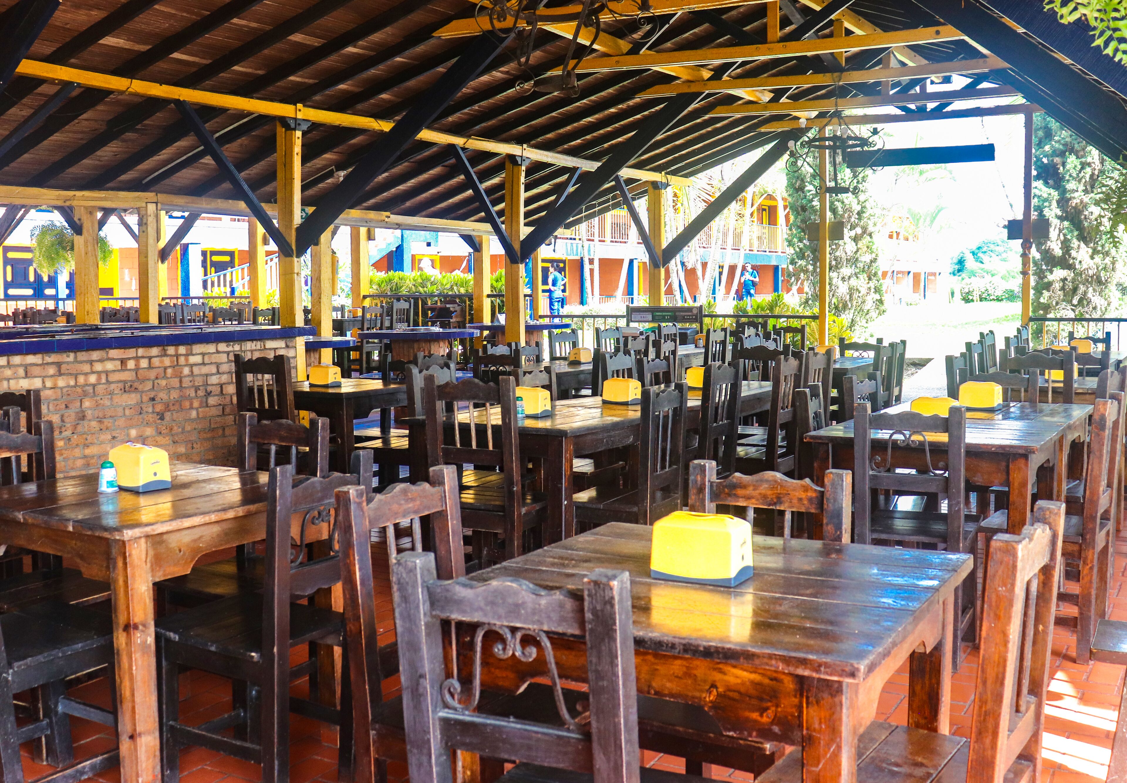 An outdoor dining area with wooden tables and chairs, a brick wall, and a covered roof structure.