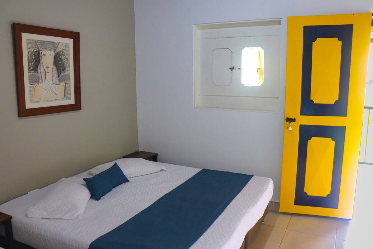 Une chambre à coucher comprenant un lit, une table de chevet, une œuvre d’art encadrée accrochée au mur, une fenêtre et une porte.