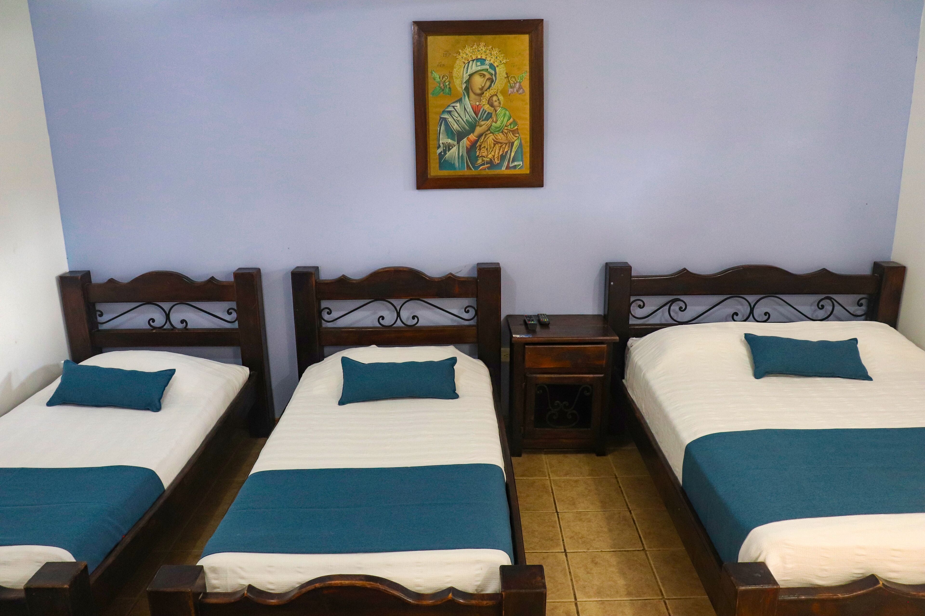 A room with three wooden beds, each with a blue bedspread and pillow. There is a small bedside table with a lamp on one side and a wall-mounted picture above the beds.
