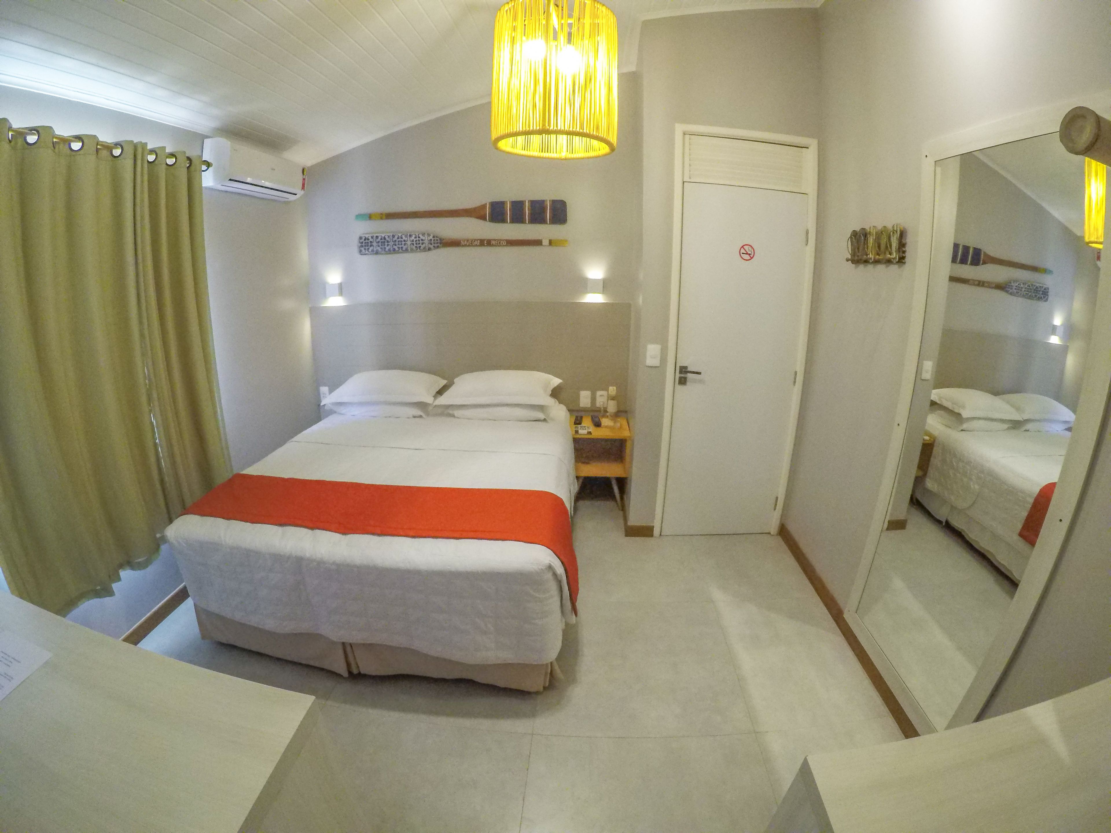 Quarto de hotel com cama, mesa de cabeceira, espelho, porta com placa e um morcego pendurado.