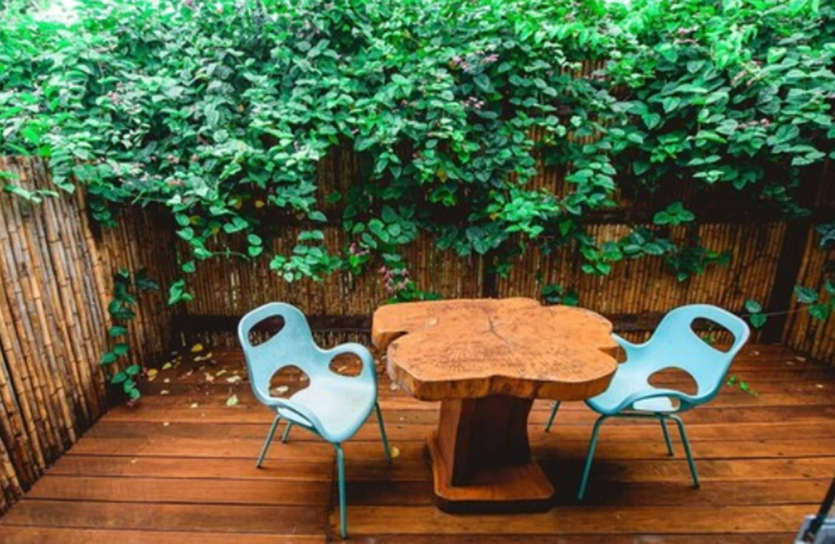 A wooden deck with a table and two chairs, surrounded by a bamboo fence and lush green foliage.