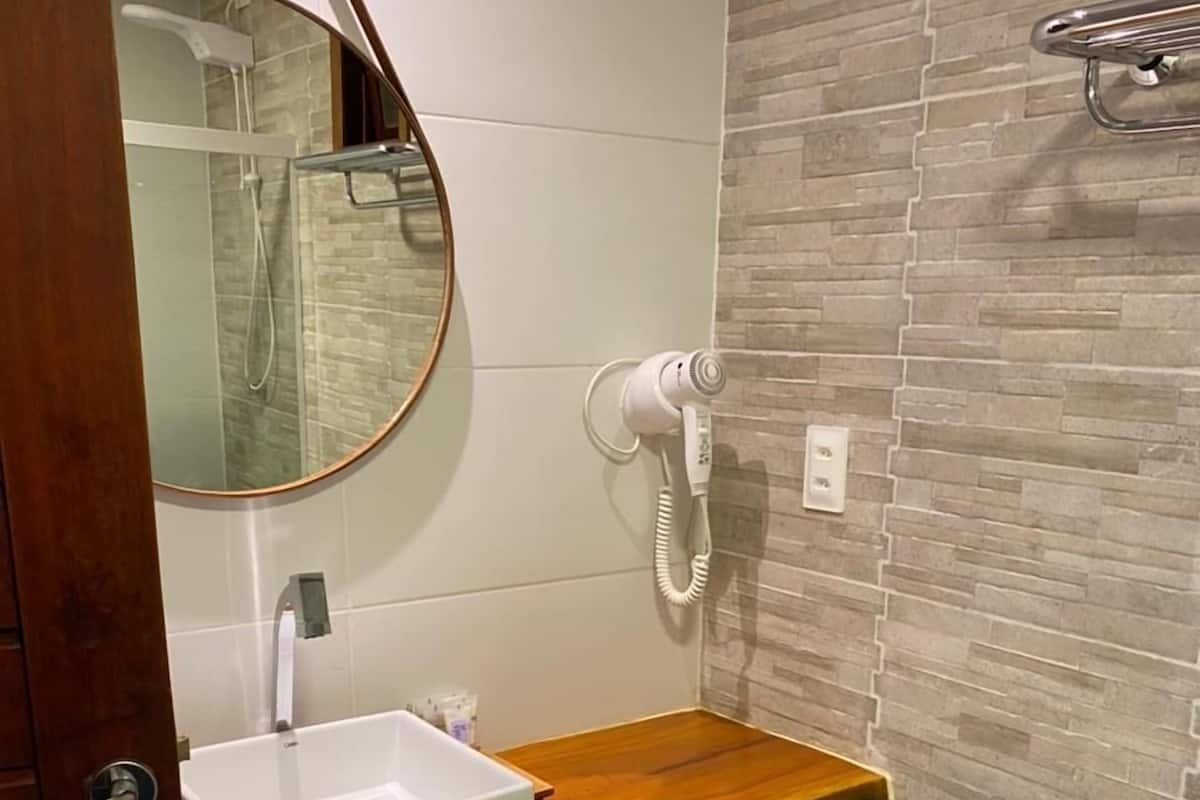 Casa de banho com toucador de madeira, lavatório branco, espelho redondo e secador de cabelo na parede.
