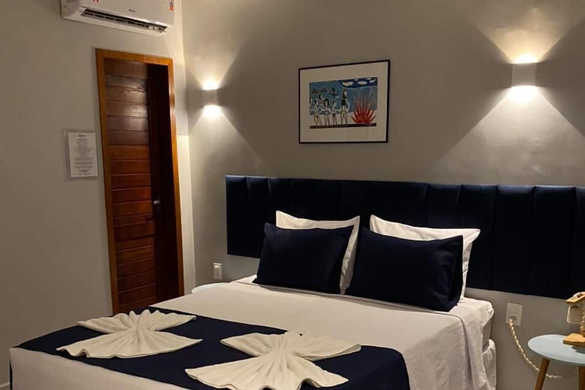 Uma cama bem arrumada com colcha branca e azul marinho, uma porta de madeira, um quadro na parede e uma mesinha de cabeceira com abajur.
