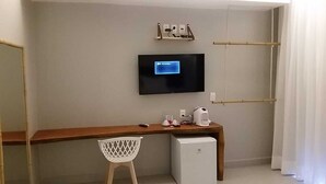 A compact hotel room with a wooden desk, a small white refrigerator, a TV mounted on the wall, and a bamboo shelf. - Pousada da Mana (Fernando de Noronha)