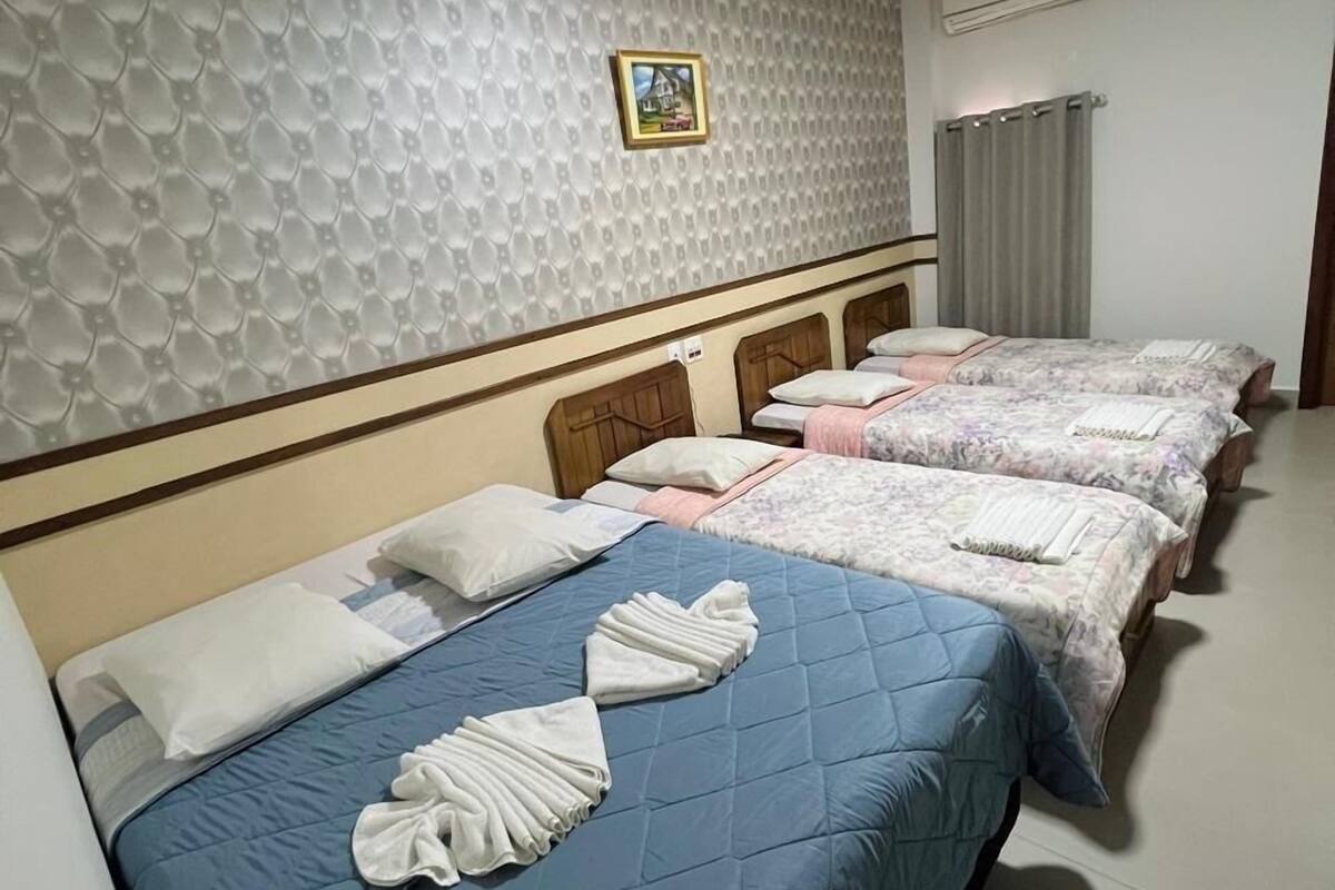 Une chambre d’hôtel avec quatre lits, chacun recouvert d’une couette bleue et agrémenté de serviettes blanches. Un climatiseur est fixé au mur, et un tableau encadré est accroché au-dessus des lits.