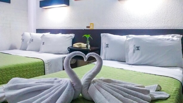Habitación de hotel con dos camas, cada una con almohadas blancas y ropa de cama verde. Hay luces fijadas en la pared sobre las camas y una planta pequeña en la mesita de noche.