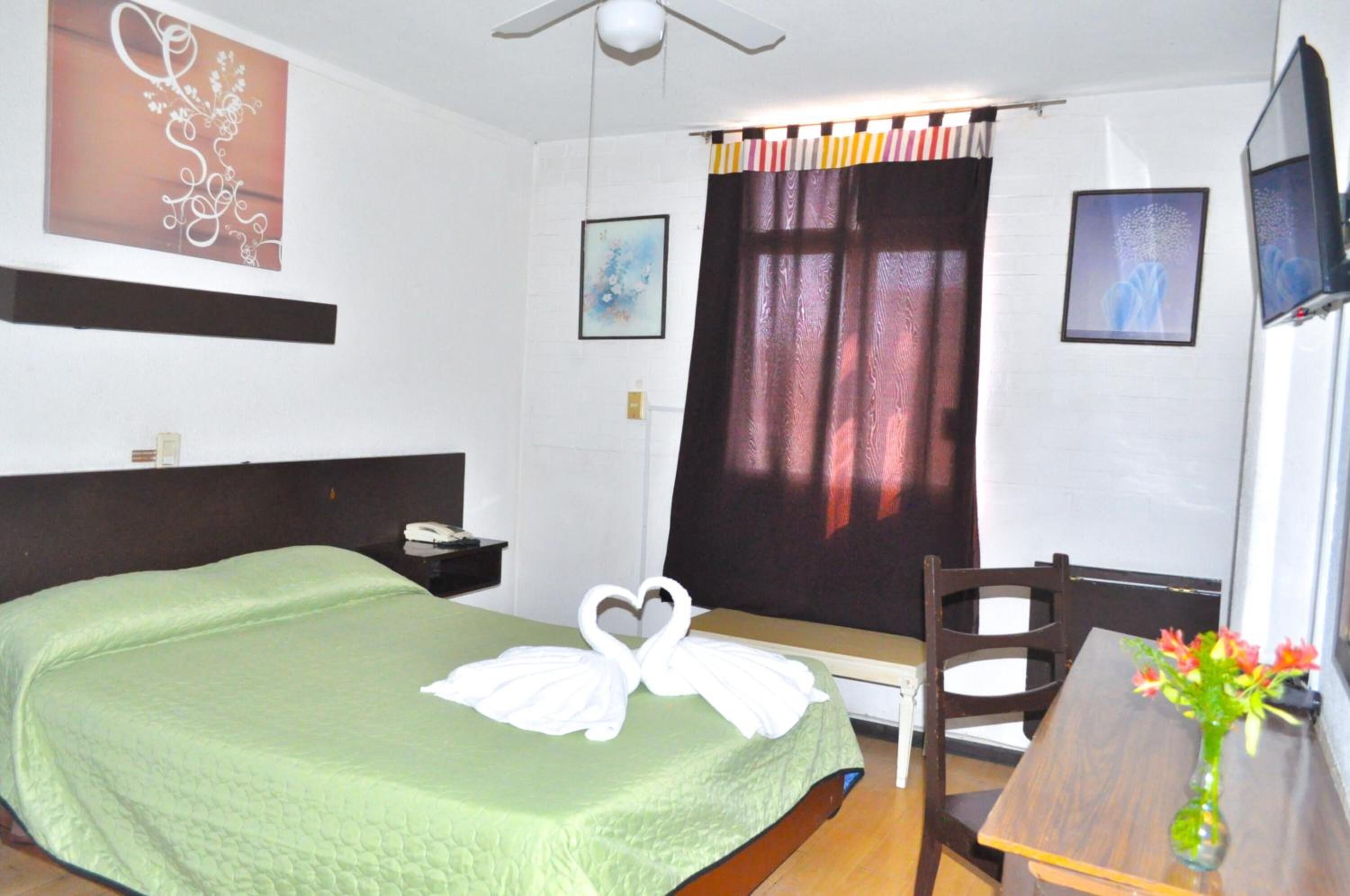 Habitación de hotel con cama, escritorio, silla, televisor y ventana con cortinas.