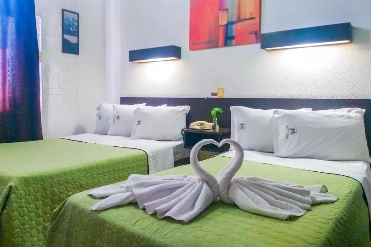 A hotel room with two beds, green bedding, white pillows, and a heart-shaped towel decoration.