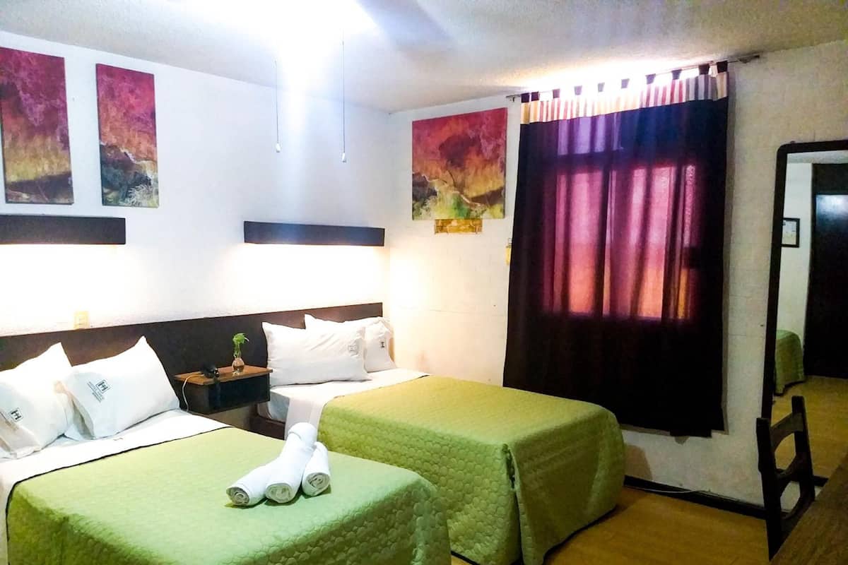 A hotel room with two beds, each with a green bedspread, white pillows, and a small table with a plant. There are two paintings on the wall and a window with purple curtains.