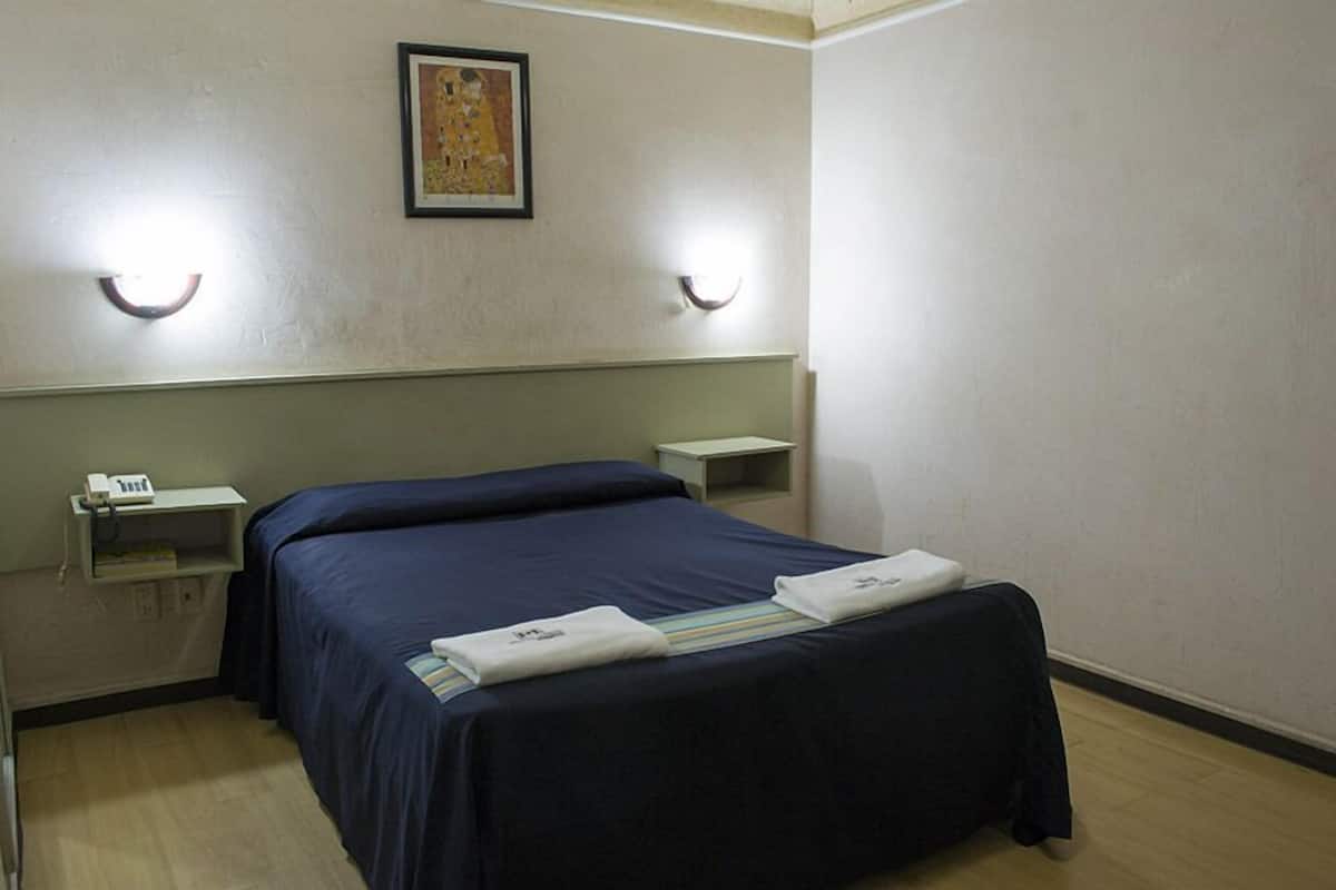 A single bed with a dark blue bedspread, two bedside tables with lamps, a small shelf with a phone, and a framed picture on the wall.