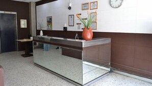 Un mostrador de recepción con separador de vidrio, un reloj, certificados enmarcados y una planta en maceta.