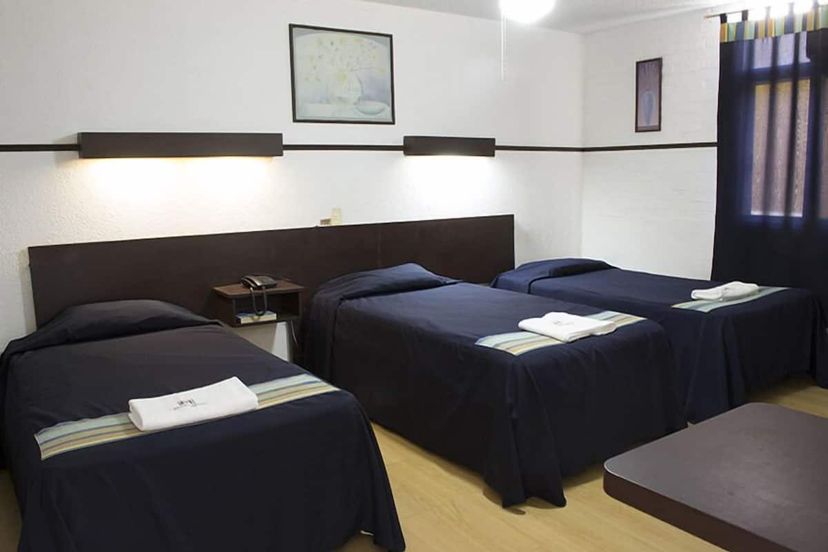 A hotel room with three single beds, a bedside table with a lamp, a phone, and a painting on the wall.