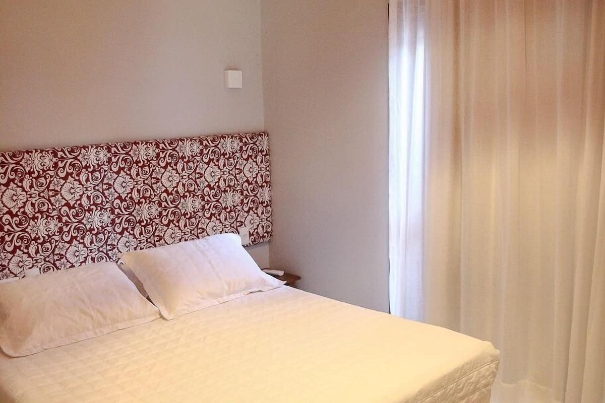 A neatly made bed with a patterned headboard, a red hanging lamp, and a white curtain.