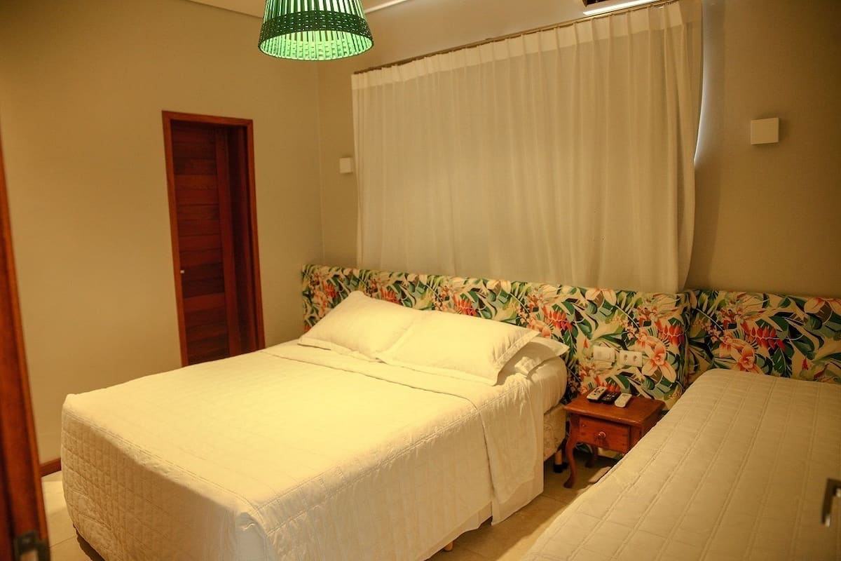 A single bed with a floral headboard, a bedside table with a remote control, and a green hanging light fixture.