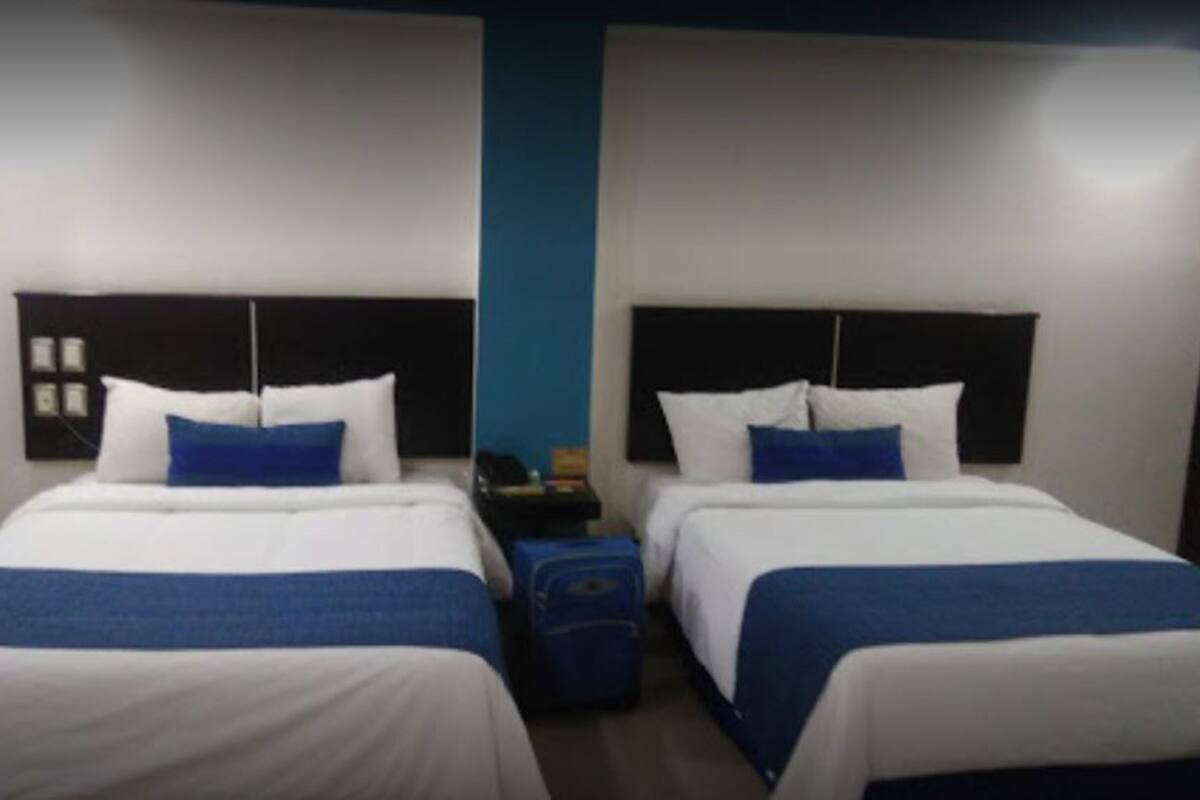 A hotel room with two beds, each with white bedding and blue accents, a blue wall, and a small table with a lamp between the beds.