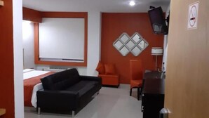 Habitación de hotel con cama, sofá negro, sillón naranja, televisor montado en la pared y puerta con cartel de no fumar.