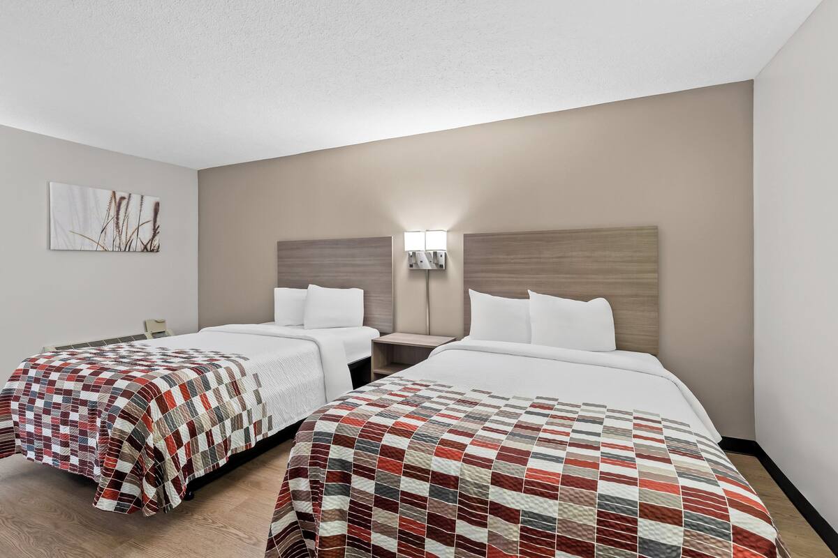 Quarto de hotel com duas camas, uma mesa de cabeceira, uma luminária na parede e um quadro.