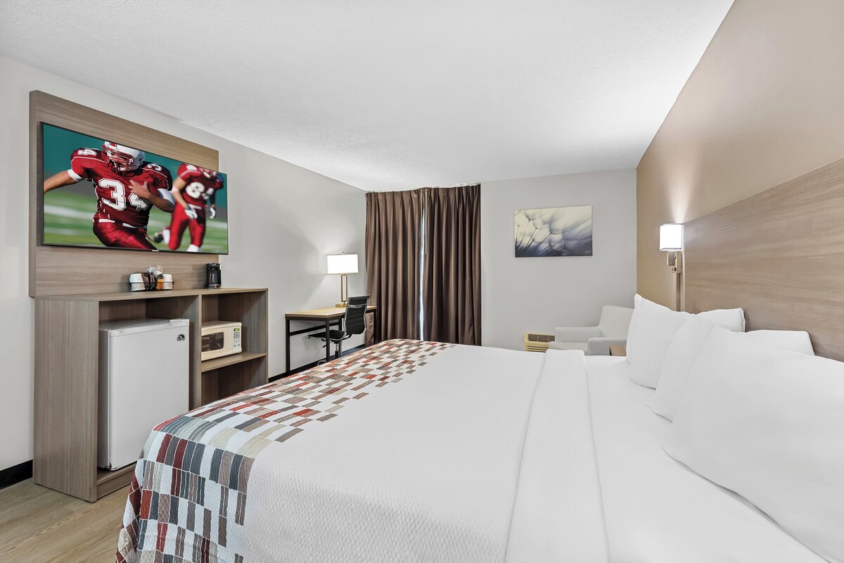 A hotel room with a large bed, a desk, a chair, a small refrigerator, a TV, and a painting on the wall.