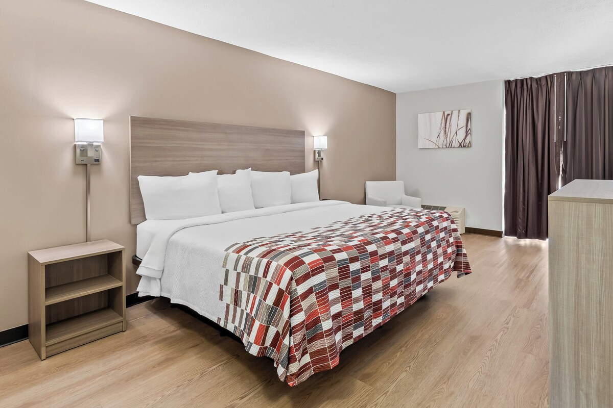 A hotel room with a bed, bedside tables, a nightstand, a chair, a desk, and a wall-mounted artwork.