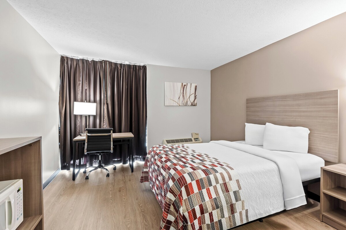 A hotel room with a bed, desk, chair, and a large window with curtains.