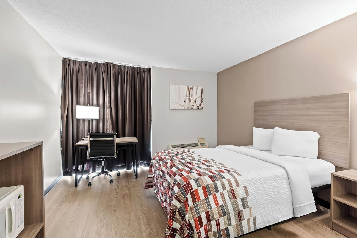 Quarto de hotel com cama, escrivaninha, cadeira e uma janela grande com cortinas.
