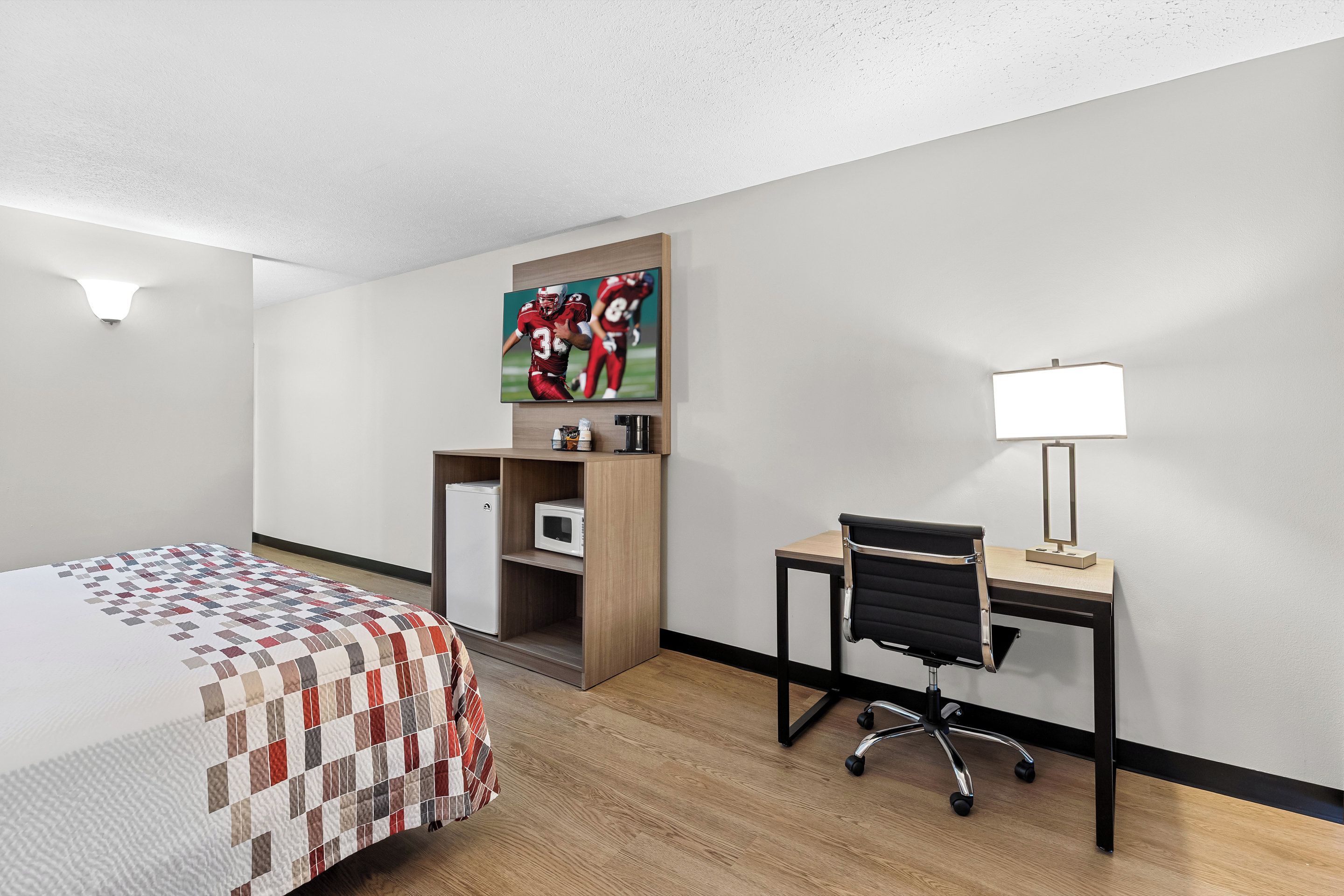 A hotel room with a bed, a desk, a chair, a TV mounted on the wall, and a lamp.