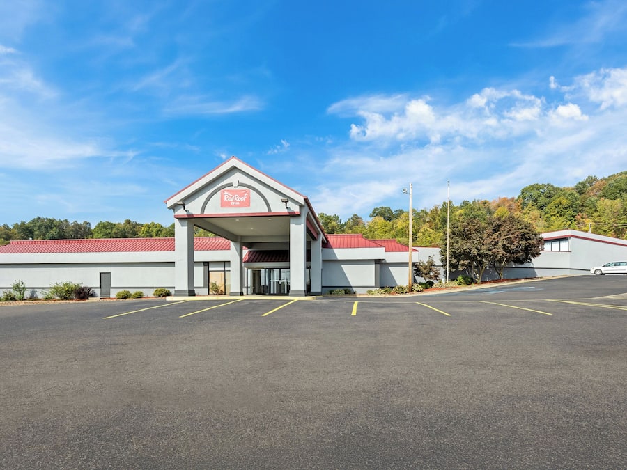 A Red Roof Inn with a red roof and white walls, a parking lot, and a hilly landscape in the background.