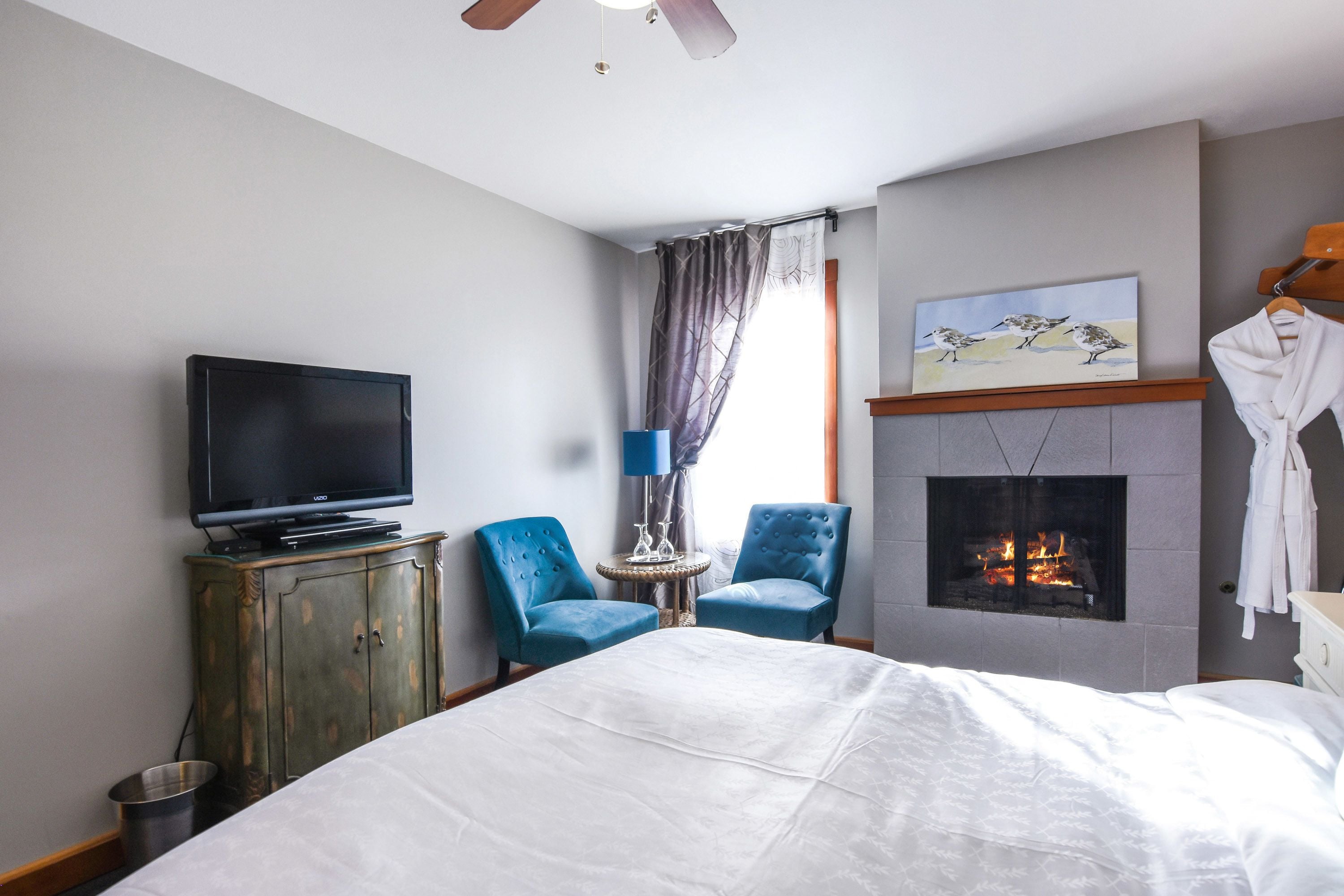 A hotel room with a bed, a television, two blue armchairs, a fireplace, and a painting on the wall.