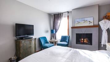 A hotel room with a bed, a television, two blue armchairs, a fireplace, and a painting on the wall.