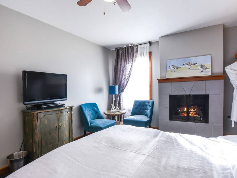 A hotel room with a bed, a television, two blue armchairs, a fireplace, and a painting on the wall.