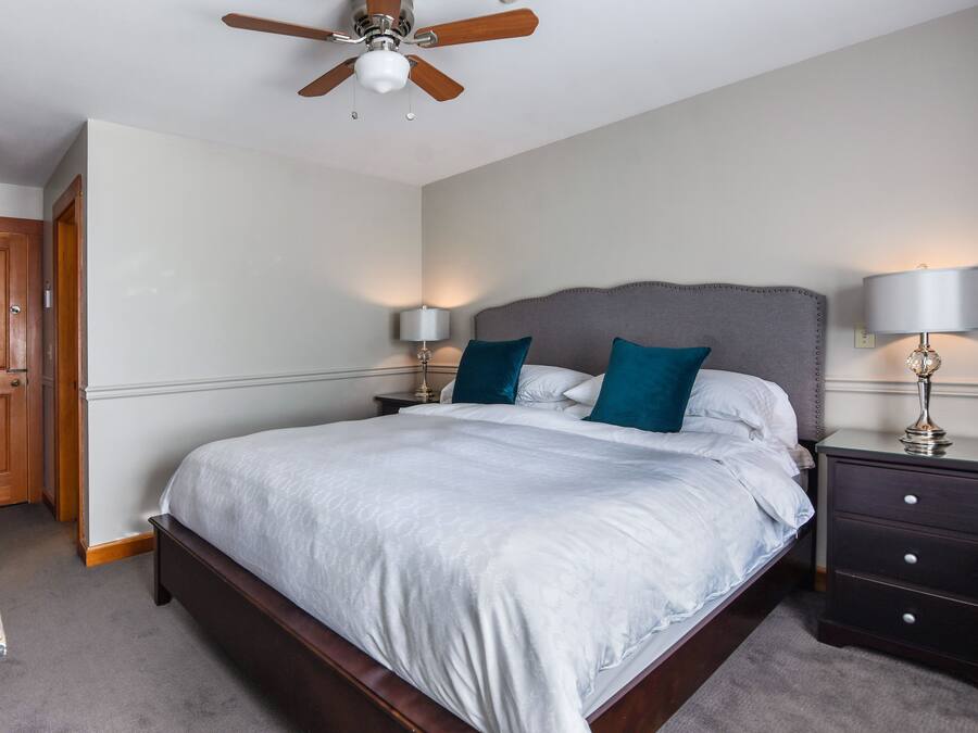 Une chambre à coucher avec un grand lit, une tête de lit en bois, deux tables de chevet avec des lampes, un ventilateur de plafond et une porte en bois.