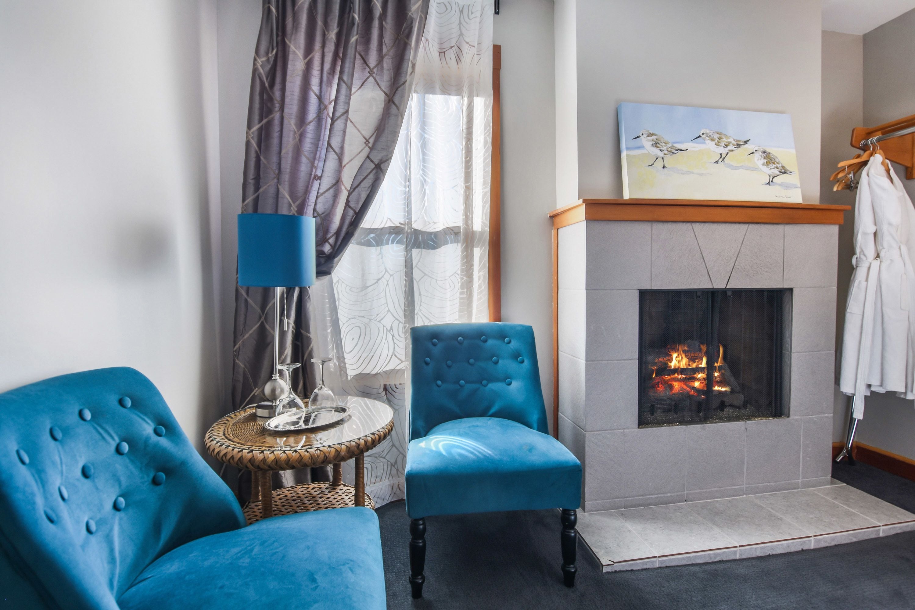 A room with a fireplace, two blue armchairs, a small round table, and a painting above the fireplace.