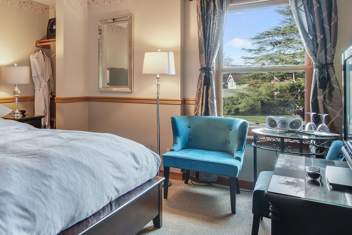 A hotel room with a large bed, two bedside tables with lamps, a chair, a small table with a glass, and a view of greenery outside.