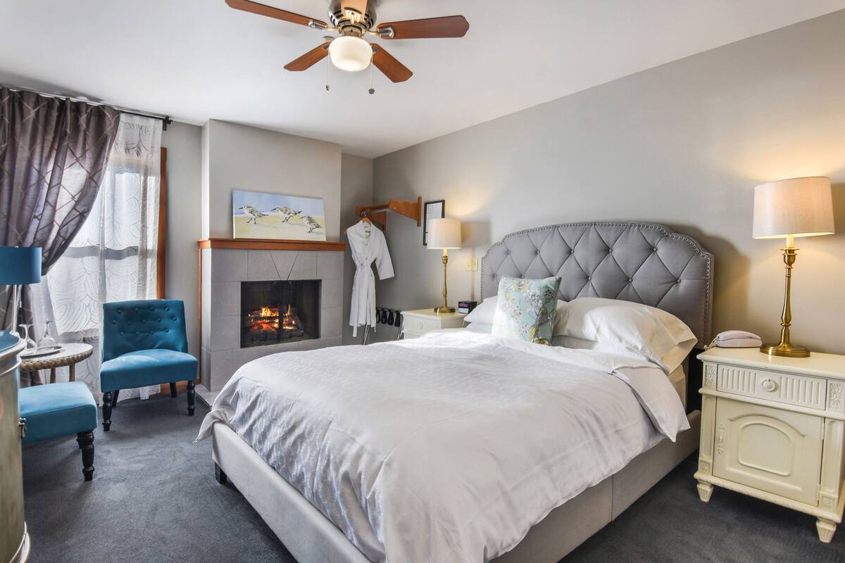 A bedroom with a large bed, a ceiling fan, a fireplace, a blue armchair, a nightstand with a lamp, and a wall-mounted robe rack.