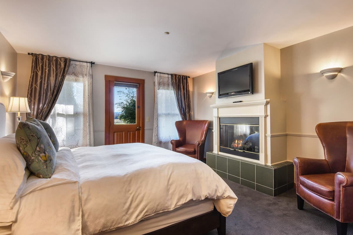 A hotel room with a large bed, two armchairs, a fireplace, a television mounted above it, and a window with curtains.