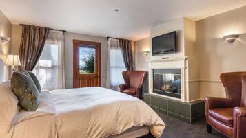 Una habitación de hotel con una cama grande, dos sillones, una chimenea, un televisor montado encima y una ventana con cortinas.