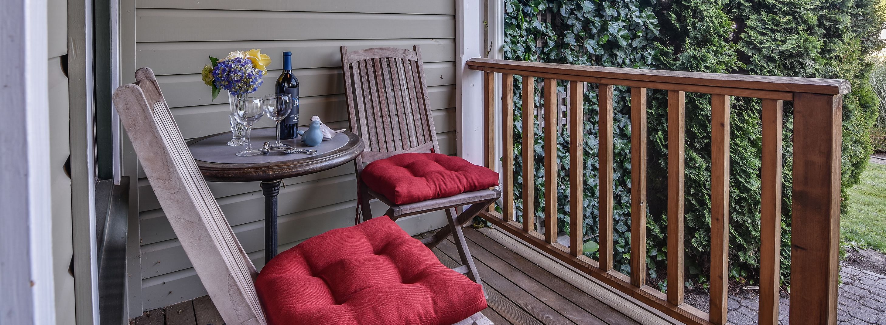 A wooden deck with a red cushioned chair and a wooden chair, a small round table with wine glasses and a bottle, and a wooden railing.