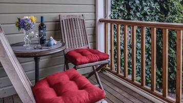 A wooden deck with a red cushioned chair and a wooden chair, a small round table with wine glasses and a bottle, and a wooden railing.