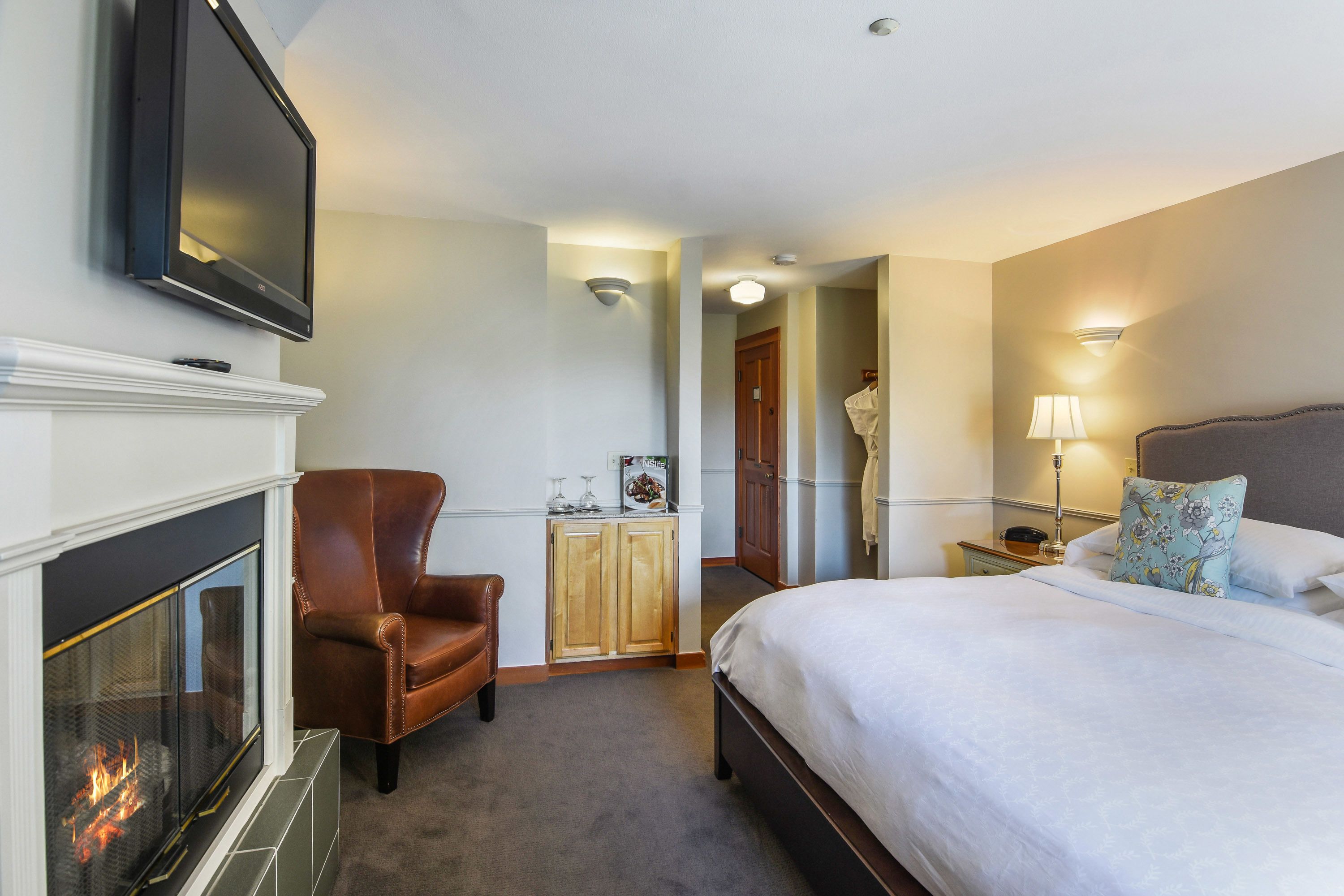 Une chambre d’hôtel avec une cheminée, un lit, un fauteuil en cuir marron, une table de chevet et un téléviseur fixé au mur.