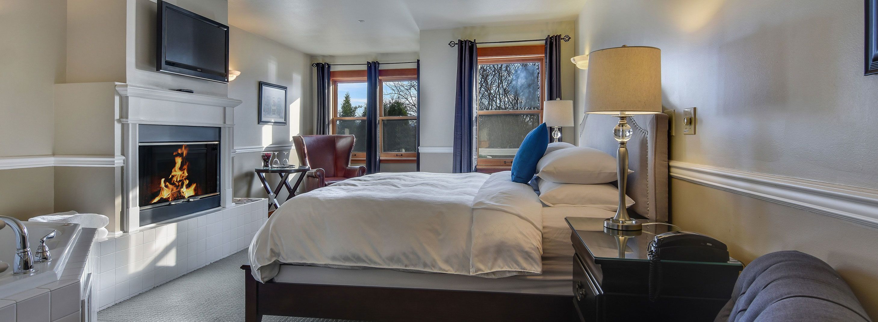 A hotel room with a fireplace, a bed, a bedside table, a lamp, and a window with curtains.