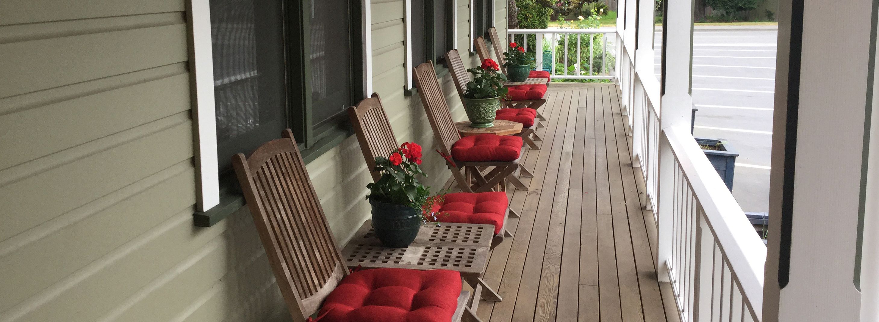 A covered porch with wooden chairs and red cushions, potted plants, and a view of the street.