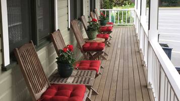 A covered porch with wooden chairs and red cushions, potted plants, and a view of the street.